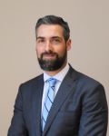 Portrait of John Drinkwater in dark gray suit with light blue tie with stripes and white collared shirt.  John has a beard, mustache and dark hair.