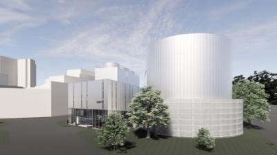 A multi level and section building, white, with some trees around it, in the forefront of the photo is a round cylinder structure
